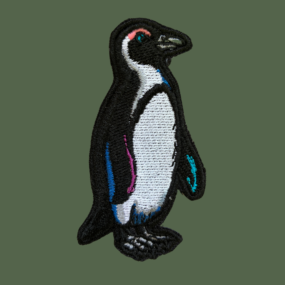 An embroidered penguin patch with a black and white body, featuring small accents of pink, blue, and teal, shown on a solid dark green background.