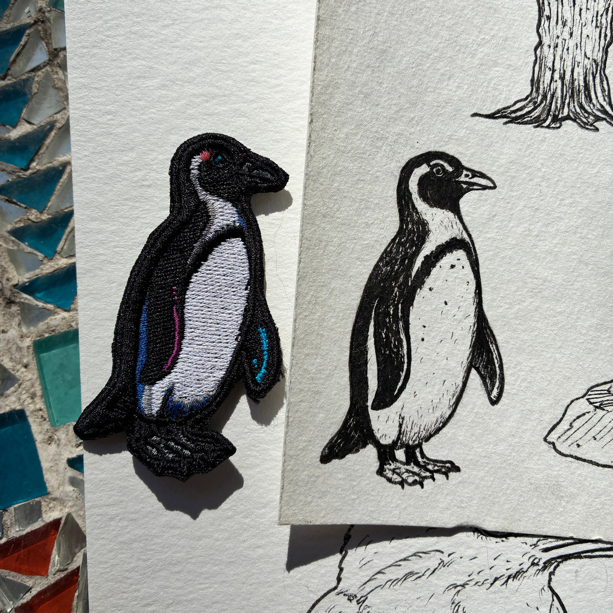 An embroidered penguin patch in black, white, and blue is shown beside a printed penguin illustration on textured paper, designed by Demi Bucklow.