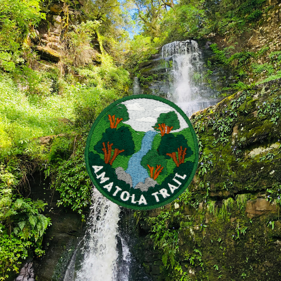 A circular embroidered patch of Amatola Trail is shown in front of a lush green waterfall and forest scene. 