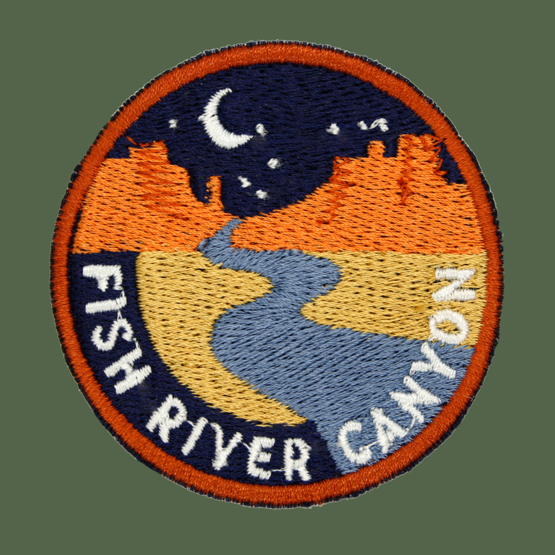 A circular embroidered patch featuring a stylized landscape with a blue river, yellow fields, orange hills, and a dark blue night sky with a white crescent moon. The patch has an orange border and white text reading “FISH RIVER CANYON” along the bottom curve.