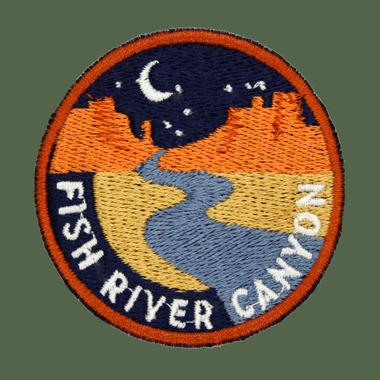 A circular embroidered patch featuring a stylized landscape with a blue river, yellow fields, orange hills, and a dark blue night sky with a white crescent moon. The patch has an orange border and white text reading “FISH RIVER CANYON” along the bottom curve.