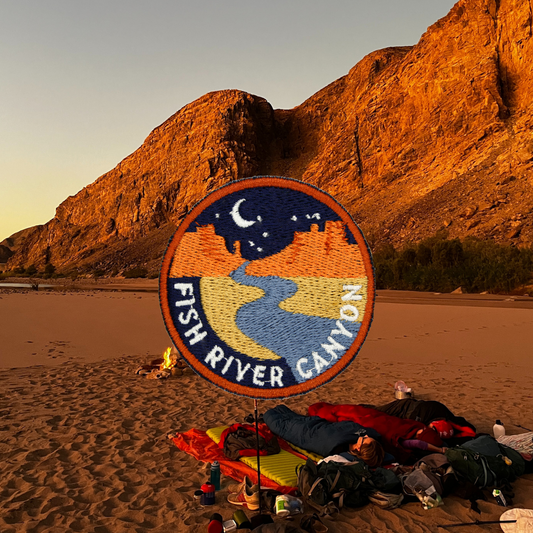 A circular embroidered patch reading 'FISH RIVER CANYON' is displayed on a stand on a sandy beach at sunset, with large rock cliffs behind it and camping gear and people sleeping underneath visible in the foreground.