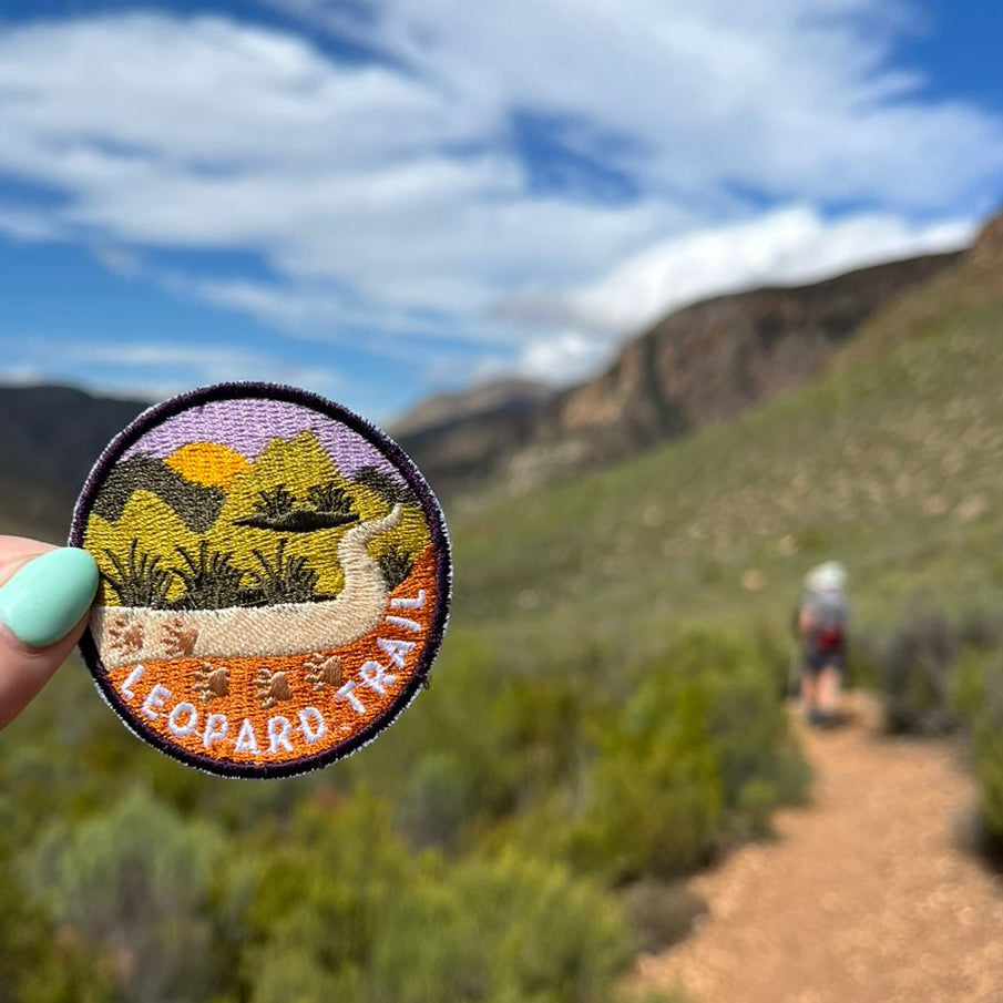 A hand holds up a circular embroidered patch reading 'LEOPARD TRAIL' against a blurred scene of the actual Leopard Trail with a dirt trail, green shrubs, and a blue sky with clouds.