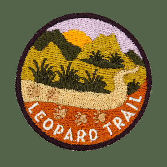 A circular embroidered patch featuring a landscape with mountains, a yellow sun, a winding beige trail, and the words 'LEOPARD TRAIL' in white.