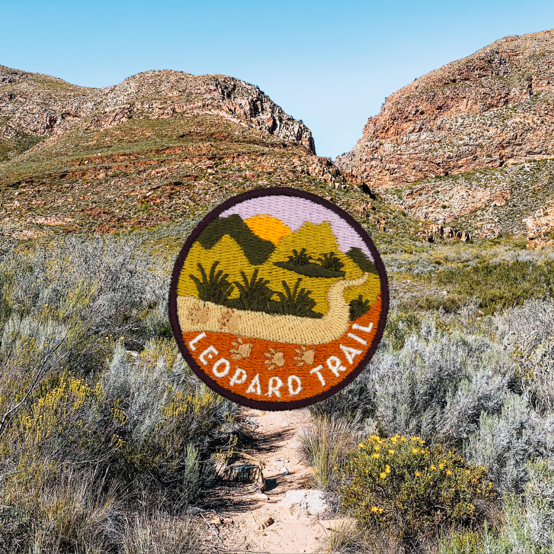 A circular embroidered patch reading 'LEOPARD TRAIL' with a landscape design, shown against a real outdoor mountain and shrub background.
