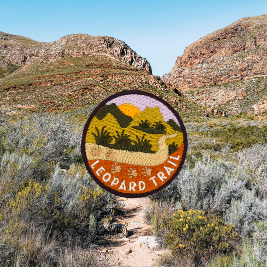 A circular embroidered patch reading 'LEOPARD TRAIL' with a landscape design, shown against a real outdoor mountain and shrub background.