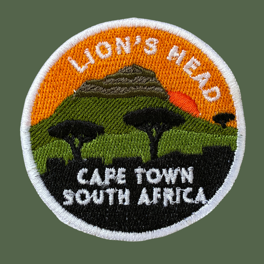A circular embroidered patch featuring a scenic landscape with the text 'LION'S HEAD' at the top and 'CAPE TOWN SOUTH AFRICA' at the bottom. The design shows an orange sky, a mountain, green hills, trees, and a black foreground, all outlined with a white stitched border.
