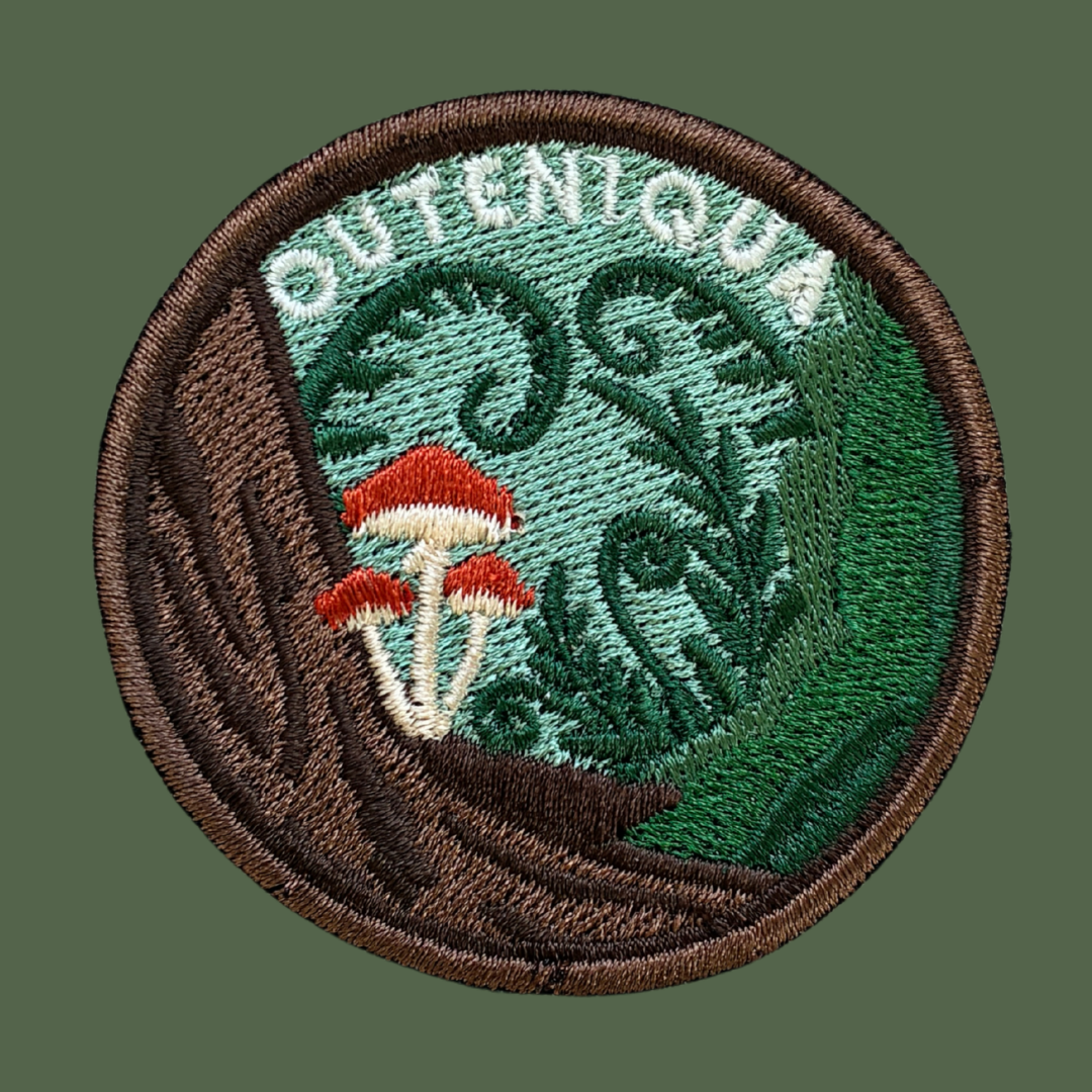 A circular embroidered patch featuring the word 'OUTENIQUA' in white lettering above a forest scene with green foliage, brown tree trunk, and three small red-and-white mushrooms, shown on a solid dark green background.