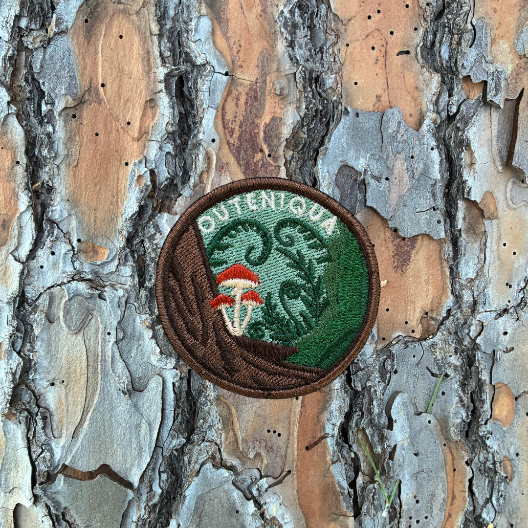 A circular embroidered patch with a brown border, featuring a green background with a stylized tree and the word 'OUTENIQUA' in white, plus a red-and-white mushroom motif, shown against textured tree bark.