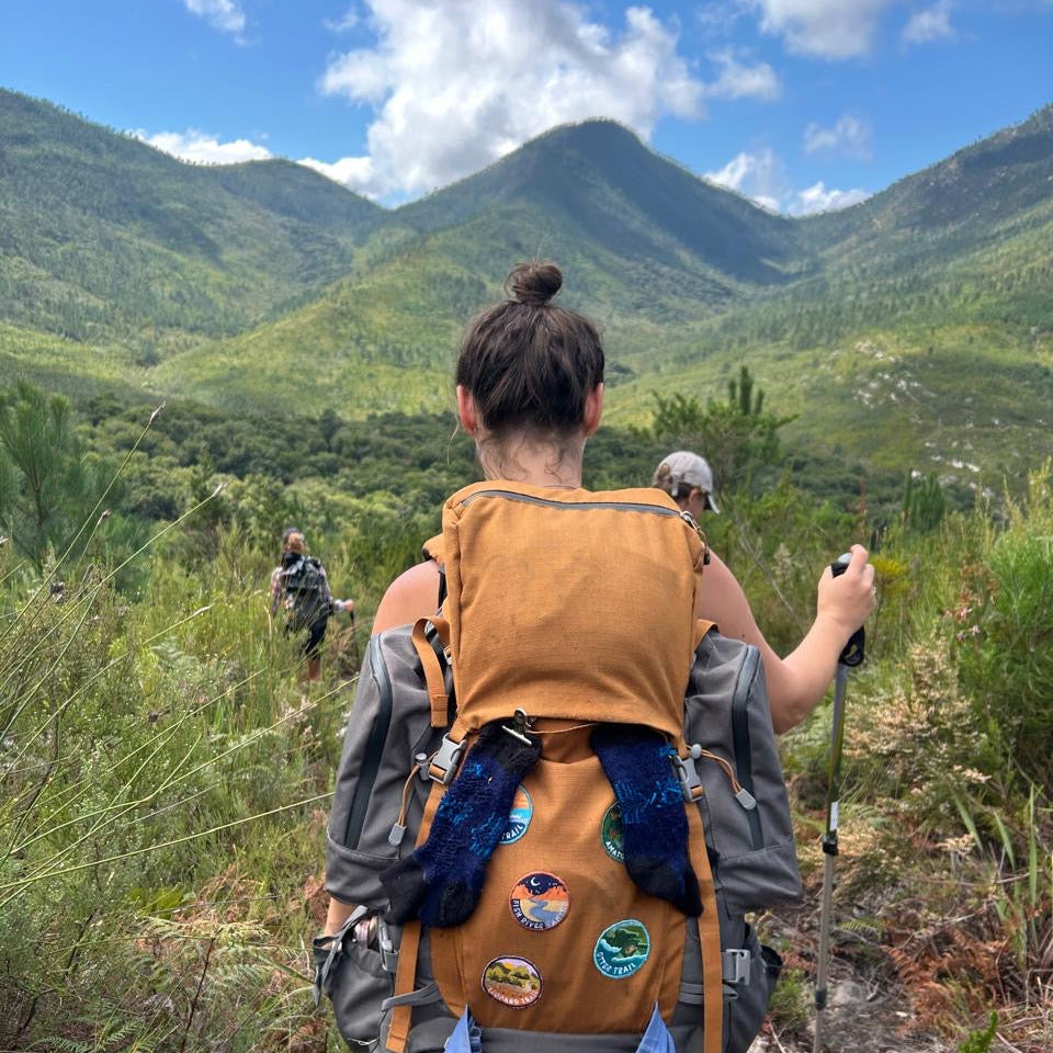 A hiker viewed from behind on Tsitsikamma Trail, wearing a large tan and gray backpack with blue accents and multiple colorful trail patches. The background shows green mountains, grassy terrain, and a blue sky with clouds.