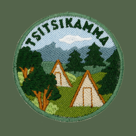 A circular embroidered patch featuring the word 'TSITSIKAMMA' arched over a landscape scene with mountains, trees, and two A-frame tents, shown on a solid dark green background.