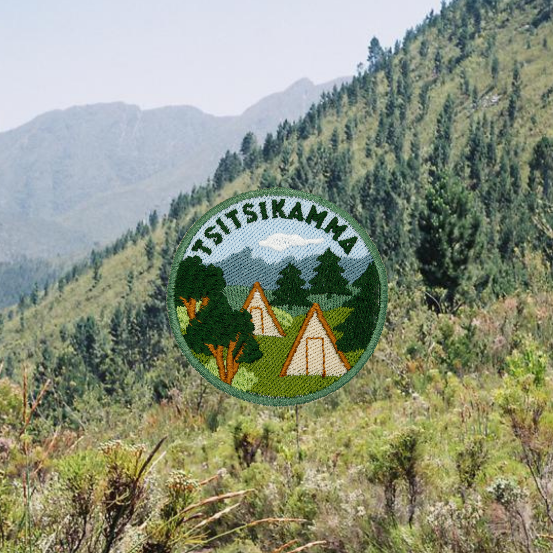 A circular embroidered patch featuring the text 'TSITSIKAMMA' above a mountain and forest scene with two A-frame cabins, shown against a blurred mountain landscape background.