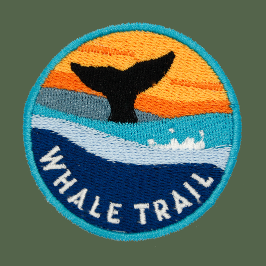A circular embroidered patch featuring a whale tail silhouette against an orange and yellow sunset, with blue ocean waves and the words “WHALE TRAIL” in white along the bottom.
