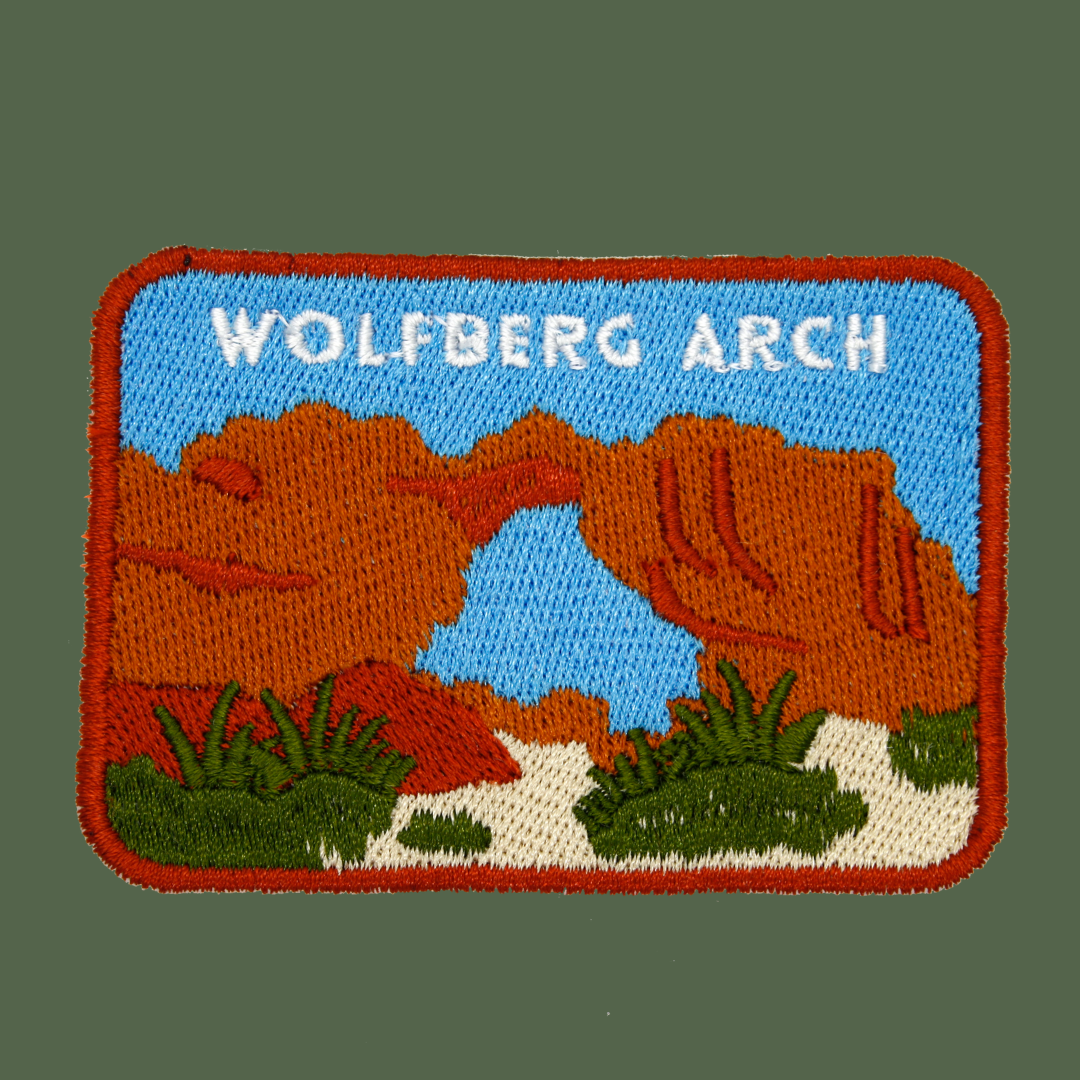An embroidered patch depicting Wolfberg Arch in a desert landscape, featuring orange rock formations, a blue sky, green shrubs, and white text reading 'WOLFBERG ARCH.'
