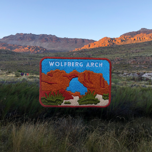 An embroidered patch depicting Wolfberg Arch in Cederberg, displayed against a real outdoor background of grassy fields and mountains at sunset.